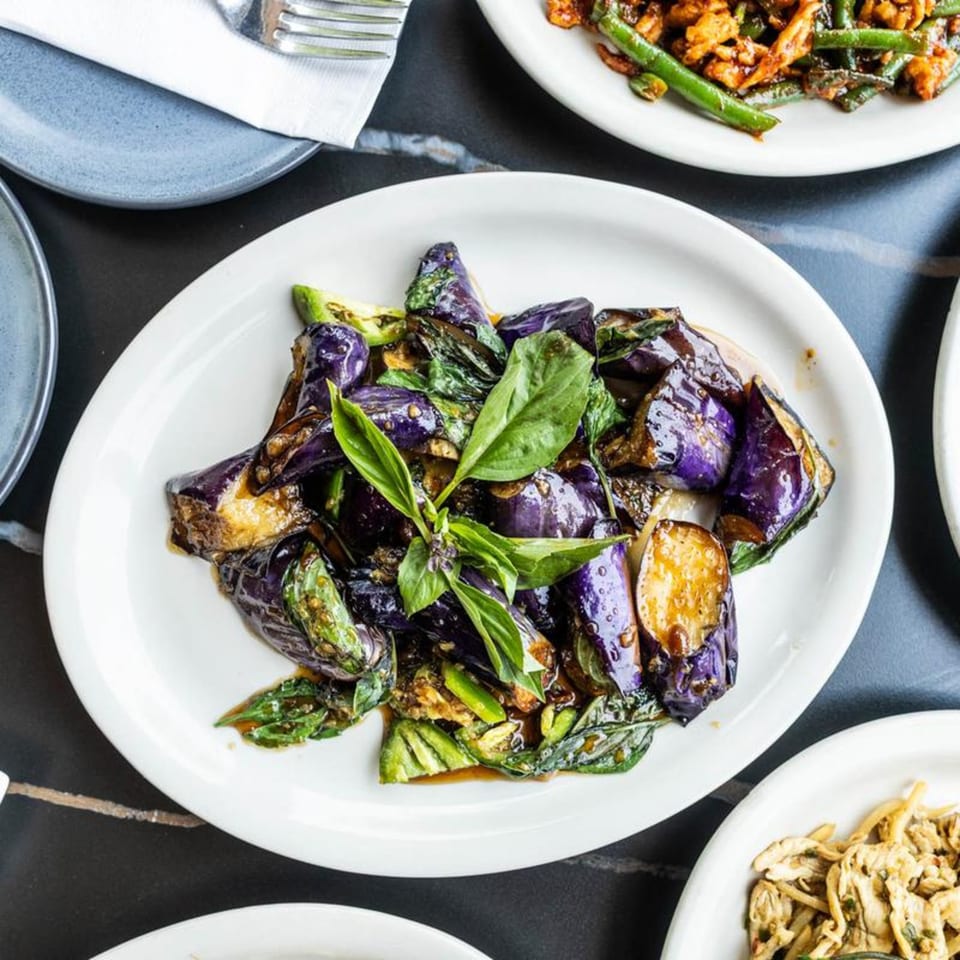 A white plate of stir-fried purple eggplant and green basil in a savory sauce.
