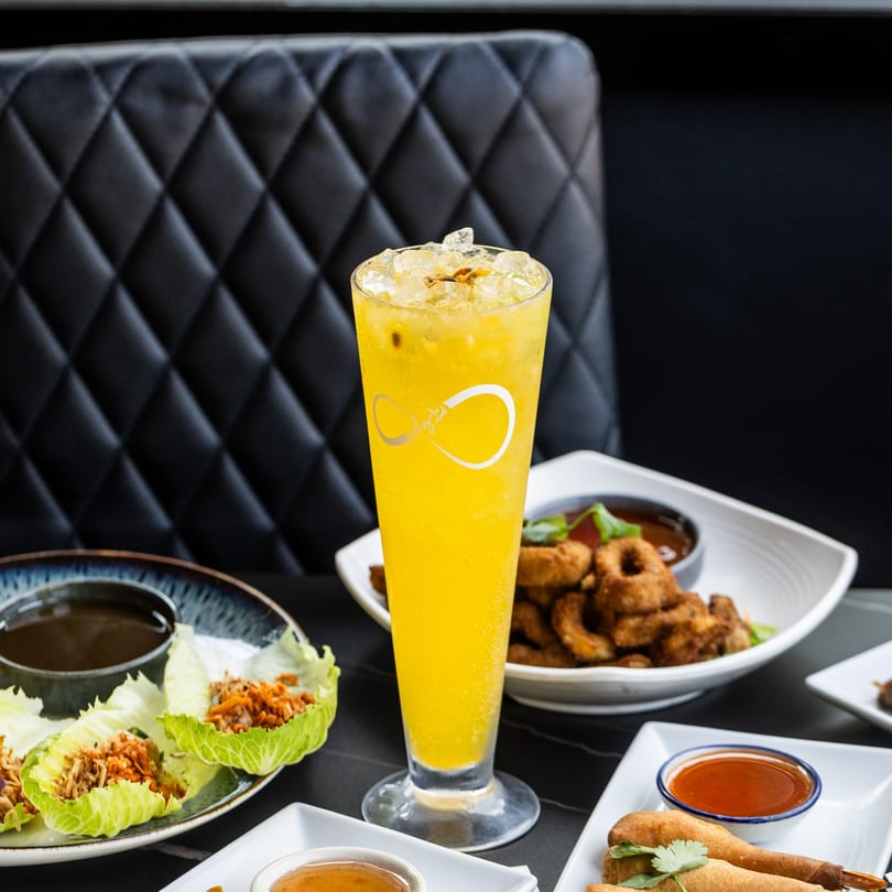 A tall glass of yellow iced drink with a logo, surrounded by various appetizers on a dark table.