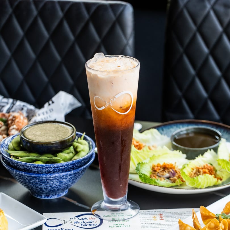 A tall glass of iced Thai tea with appetizers, including edamame and lettuce wraps, on a dark table.