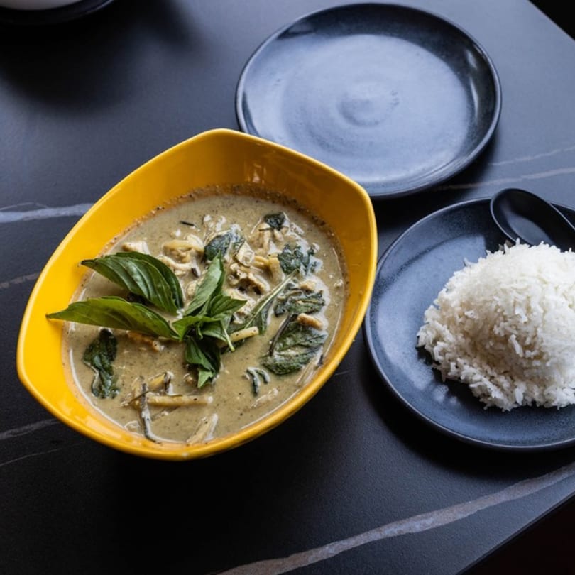 Green curry with chicken and basil in a yellow bowl, served with white rice on a dark plate.