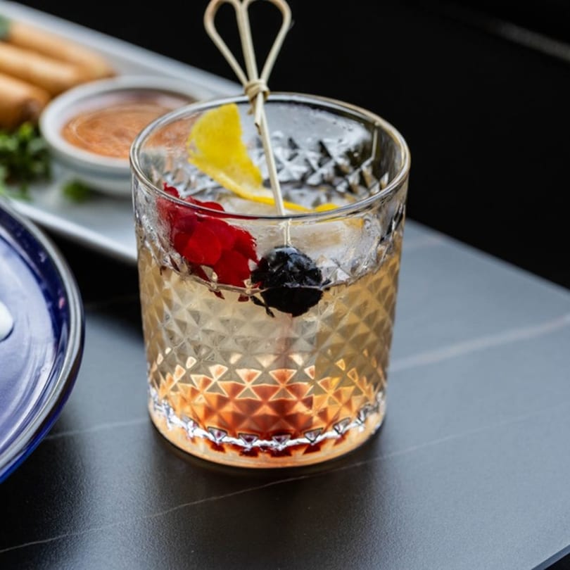 A patterned glass of amber cocktail with ice, lemon, and cherries, next to appetizers.
