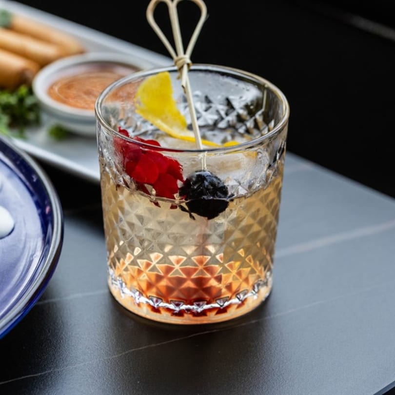 A cocktail with ice, lemon, and cherries in a textured glass, next to spring rolls.