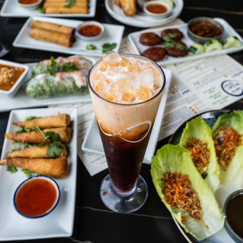 A tall glass of iced Thai tea surrounded by various Thai appetizers and dipping sauces.