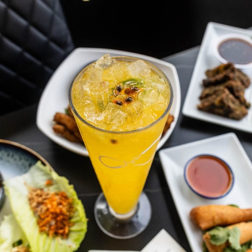 A vibrant yellow passion fruit drink with ice and mint, surrounded by various appetizers on a dark table.