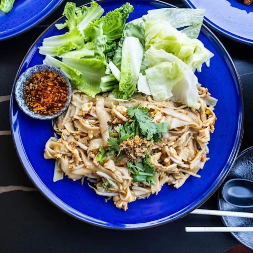 Chicken pad see ew with fresh lettuce and a side of chili sauce on a blue plate.