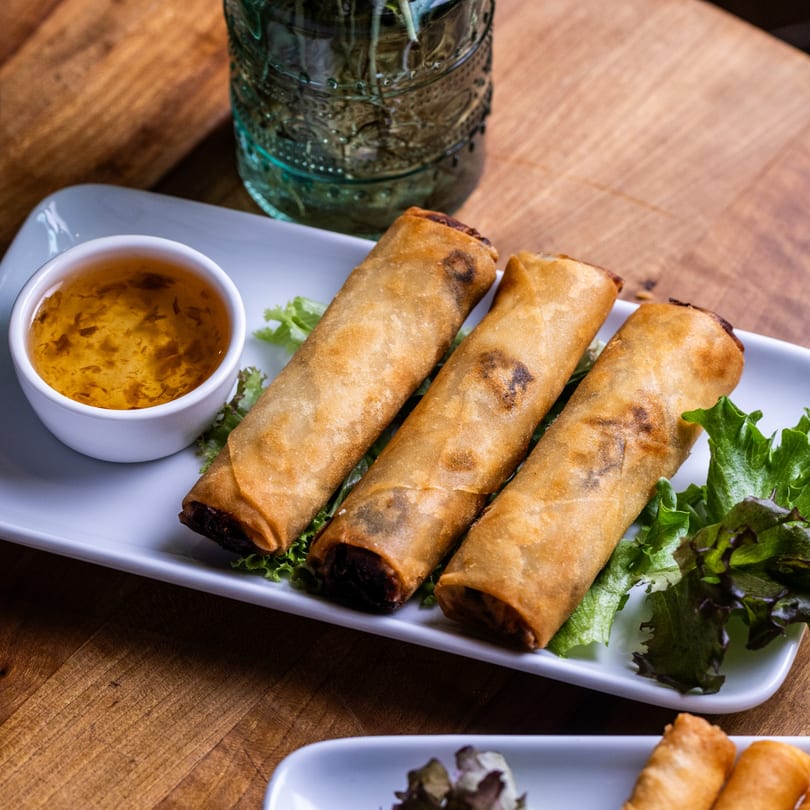 Three crispy spring rolls on lettuce with a side of dipping sauce, on a white plate.