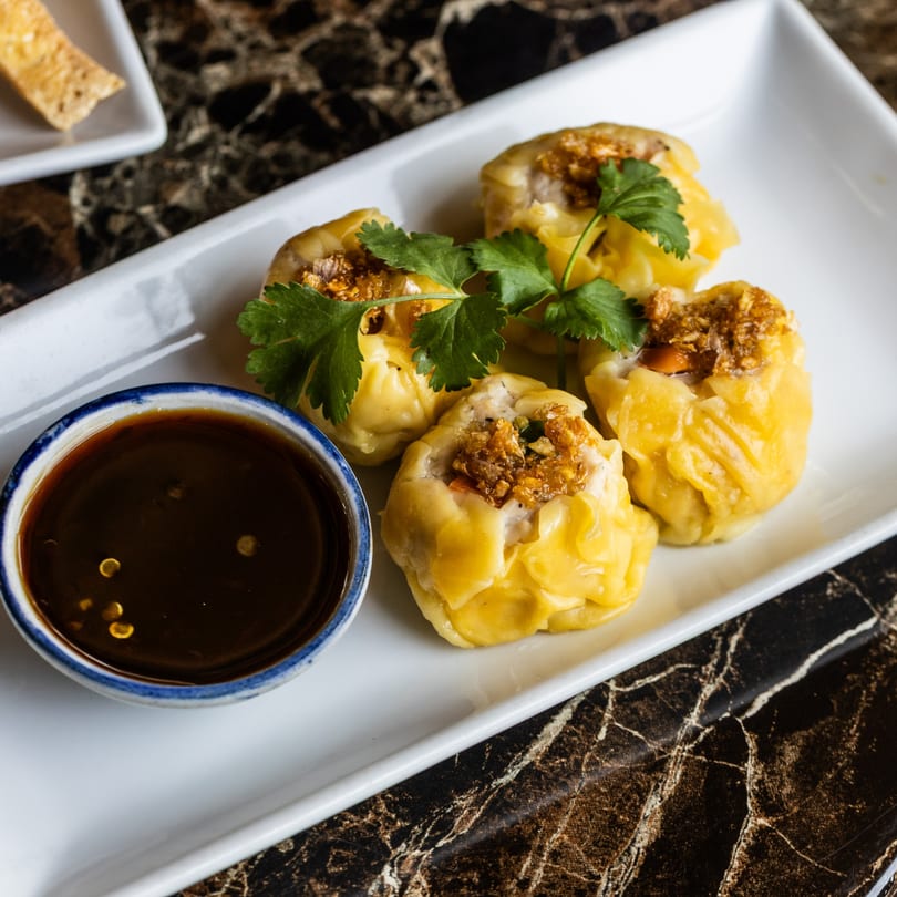 Four steamed dumplings with crispy garlic and cilantro, served with a side of dark dipping sauce.