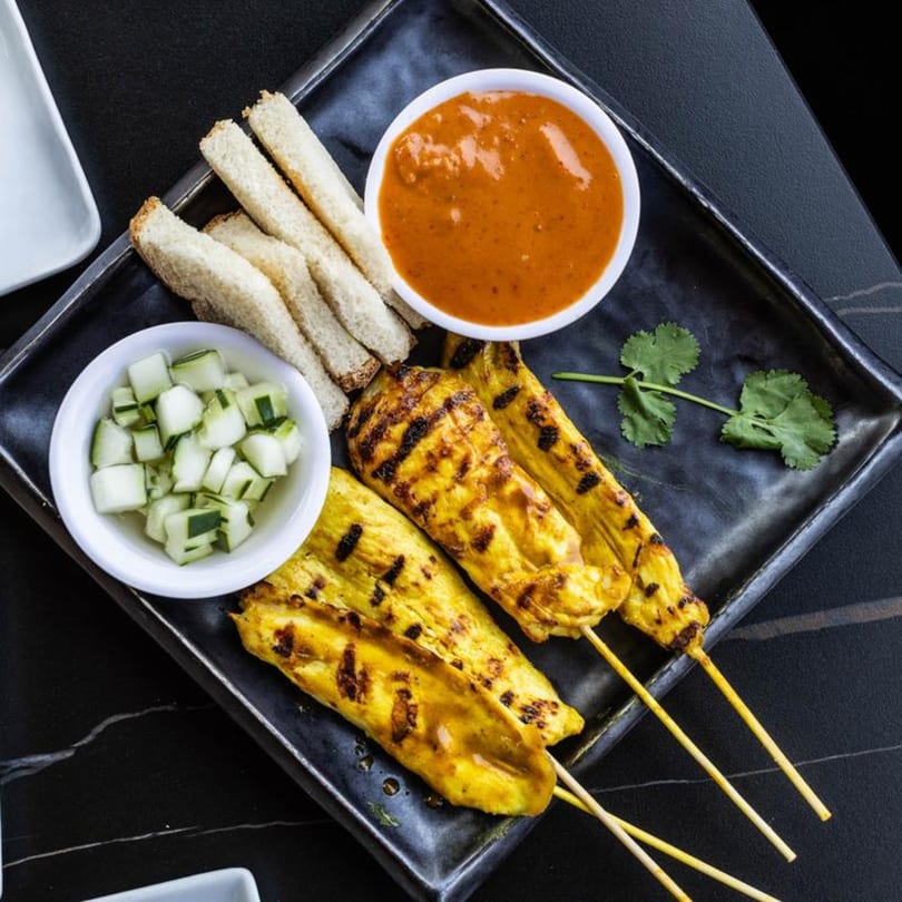 Grilled chicken satay skewers with peanut sauce, cucumber salad, and toast on a black plate.